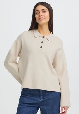 Vrouw met lang donker haar, dragend een beige trui met lange mouwen en kraag met drie knopen en een blauwe spijkerbroek, staand tegen een effen achtergrond.