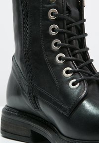 Gros plan sur une botte en cuir noir à lacets, avec œillets métalliques et fermeture éclair latérale sur une semelle cousue.