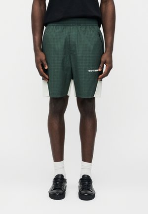 Mann trägt dunkelgrüne karierten Sportshorts mit weißen Seitenpaneelen, weiße Socken, schwarze Turnschuhe und ein schwarzes Shirt, steht aufrecht.