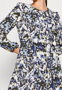 Mekko pitkähihaisella mallilla, jossa on monivärinen ikat-kuvio sinisen, vihreän ja valkoisen sävyissä. Mukana on vyötärölle istuva malli vyön kanssa.