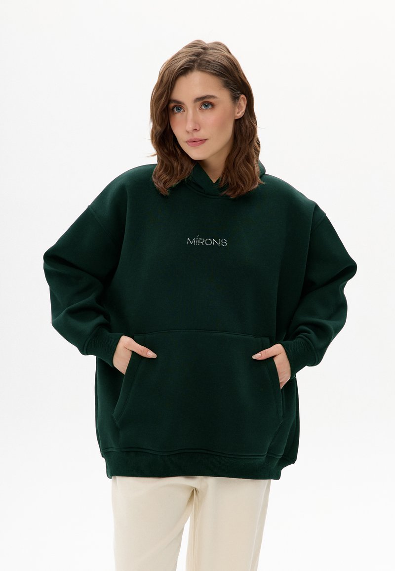 Vrouw met golvend bruin haar, gekleed in een donker groene oversized hoodie met zakken aan de voorkant en lichtbeige broek, staand tegen een witte achtergrond.