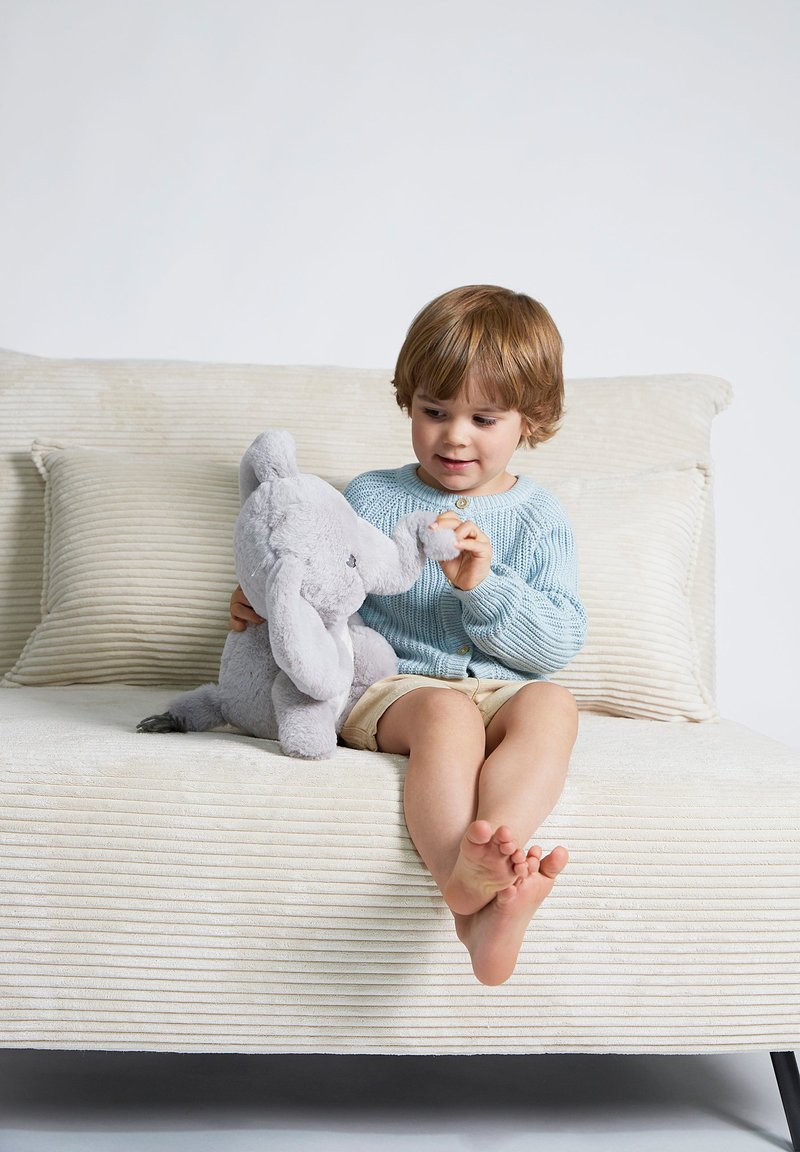 Bambino che tiene un morbido peluche di un elefante grigio chiaro, indossando un maglione azzurro e pantaloni corti beige, seduto su un divano beige con texture.