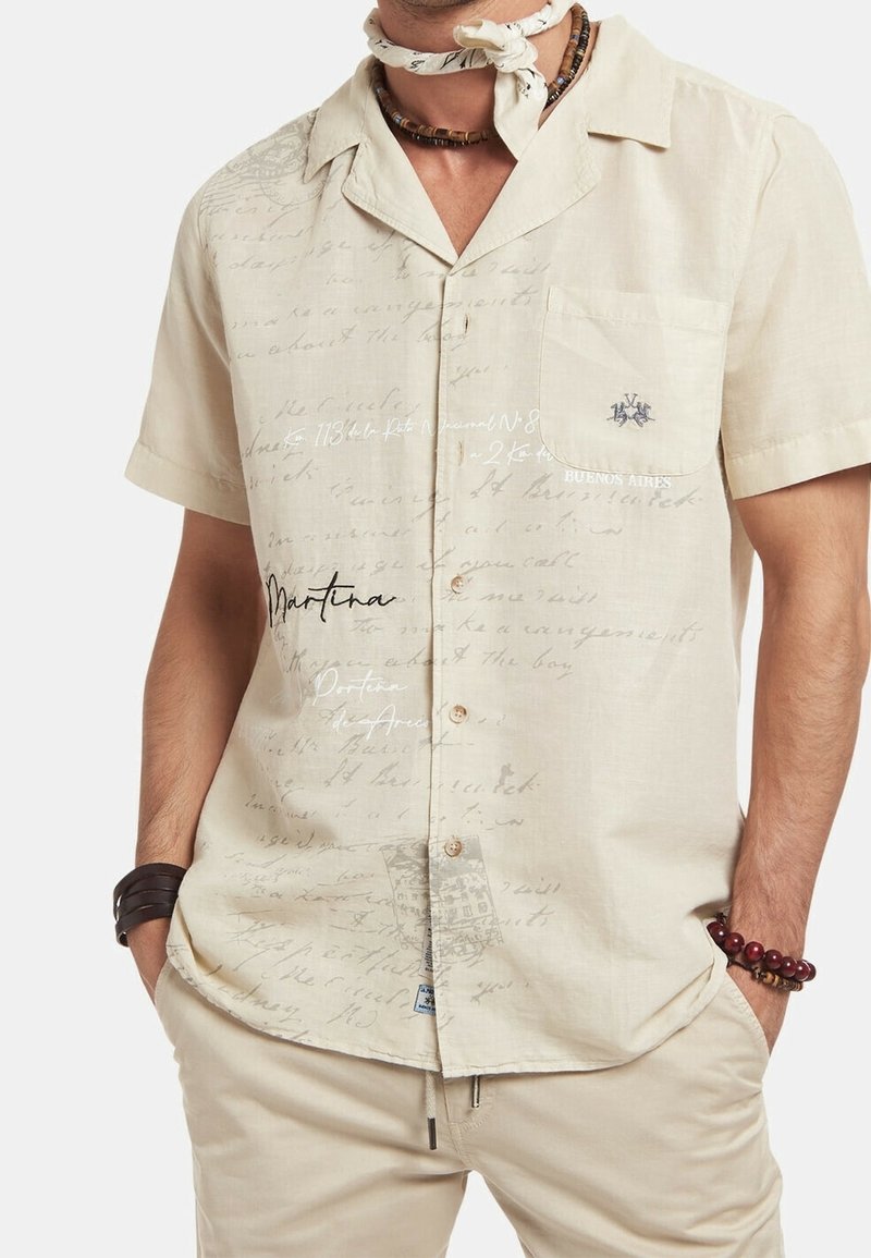 Světlý béžový krátký rukáv košile s ručně psaným vzorem textu, náprsní kapsou a knoflíkovým zapínáním. Detail textu a emblému na látce.