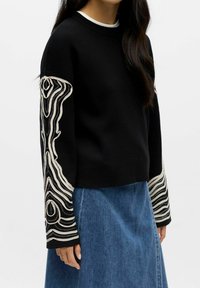 Svart genser med lange ermer, preget av intrikat, kremfarget abstrakt broderi. Kombinert med en blå denimskjørt.