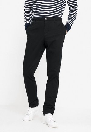 Personne portant un pantalon slim noir, des baskets blanches et un haut à manches longues rayé bleu marine et blanc avec les mains dans les poches.