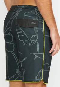 Pantaloncini da bagno Volcom neri con disegni bianchi di piante; caratterizzati da tasca laterale con pattina e cuciture a contrasto verdi all'orlo.