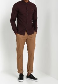 Hombre con camisa de botones color burdeos oscuro, pantalones caqui remangados y zapatillas negras con suelas blancas, de pie con las manos en los bolsillos.