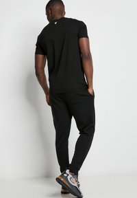 Camiseta negra de manga corta con un pequeño logo en la parte posterior, combinada con pantalones jogger negros y zapatillas deportivas grises con acentos naranjas.