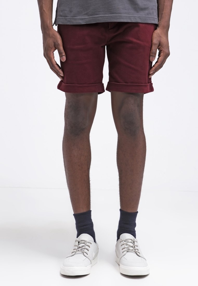 Homme portant un short bordeaux remonté, un t-shirt gris, des chaussettes noires et des baskets blanches, debout devant un fond blanc.
