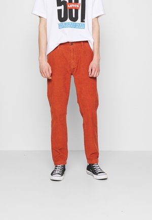 Persoon die roestkleurige broek, een wit Levi's grafisch t-shirt en zwart-witte sneakers draagt, staat tegen een effen lichte achtergrond.
