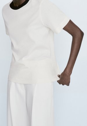 Persoon draagt een wit shirt met korte mouwen en een zwarte kraag en witte broek, trekt het shirt iets op bij de zoom tegen een lichte achtergrond.