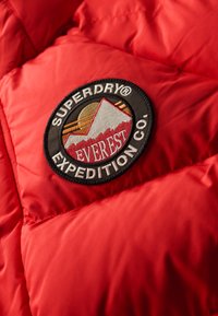 Rød pufferjakke med kviltet tekstur. Har et cirkulært sort mærke med "Superdry® Everest Expedition Co." og bjergbillede.