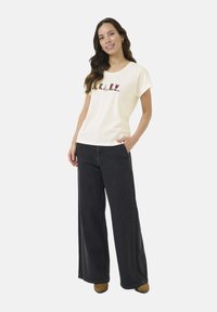 T-shirt à manches courtes de couleur crème avec des motifs de bottes colorées brodés, associé à un jean ample gris foncé.