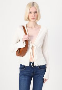Biely textúrovaný kardigán s viazaním, s volánovými rukávmi a riaseným lemom, skombinovaný s tmavomodrými džínsy a hnedou crossbody taškou.