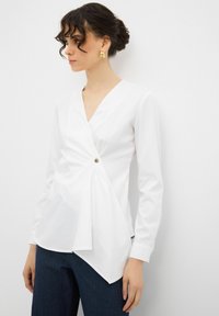 Blusa bianca avvolgente con maniche lunghe, scollatura a V e dettaglio con bottone laterale. Presenta un tessuto drappeggiato che crea un orlo asimmetrico.