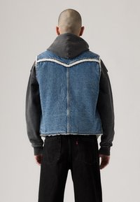 Denimvest med frynset kant og kontrasterende lys blå farve, båret over en grå hættejakke og sorte bukser, set fra bagved.