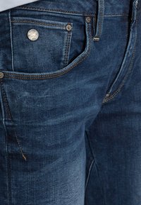 Dark blue denim jeans with a slightly faded texture, featuring orange stitching, a metal button, and a small front pocket.
