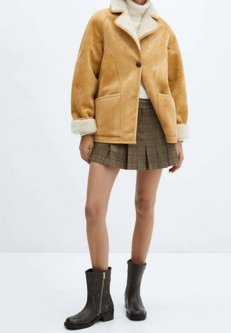 Personne portant une veste en peau retournée beige, un pull à col roulé blanc, une jupe plissée à carreaux marron et des bottes noires mi-mollets, debout devant un fond blanc.