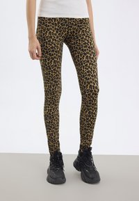 Person klädd i bruna och svarta leopardmönstrade leggings med svarta plattformssneakers på ett grått golv och vit bakgrund.