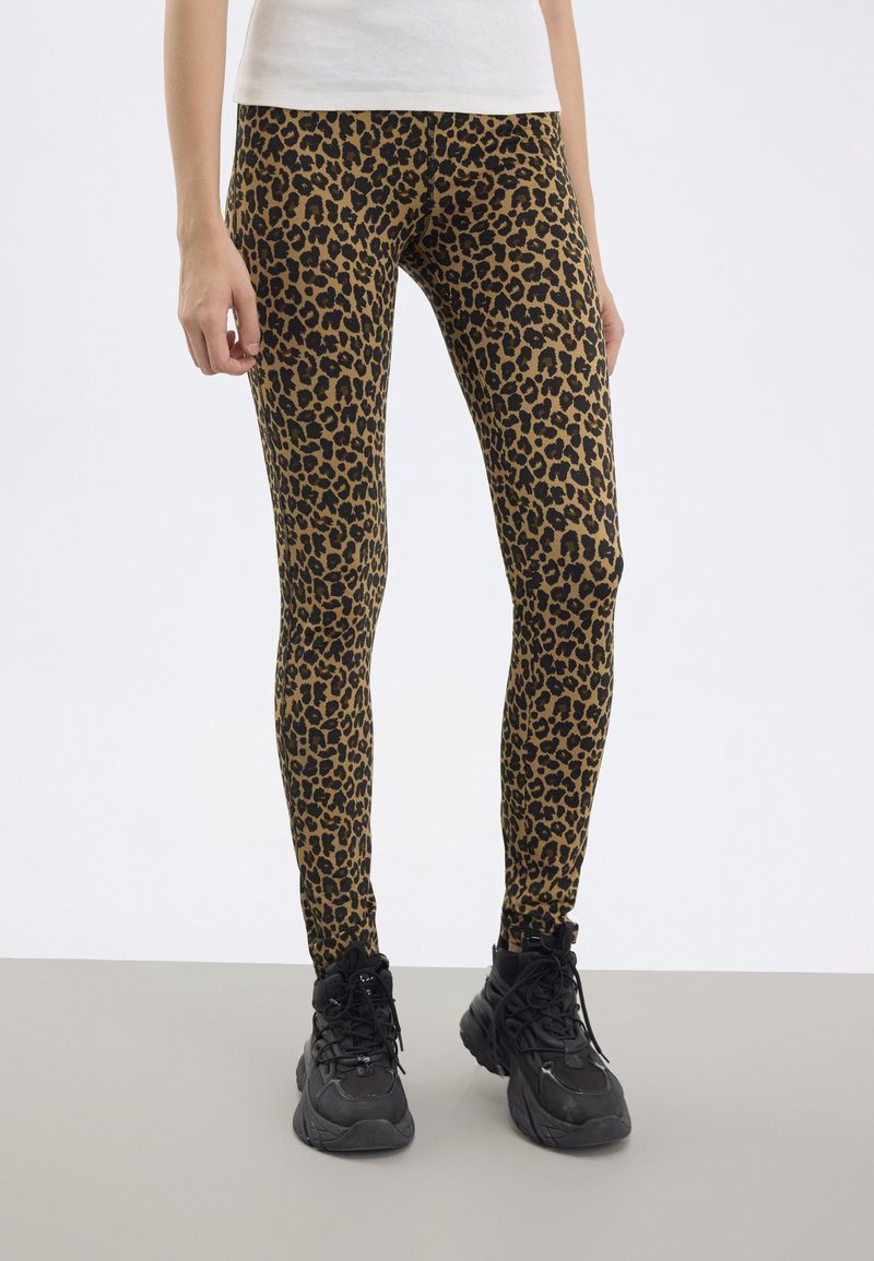 Person klädd i bruna och svarta leopardmönstrade leggings med svarta plattformssneakers på ett grått golv och vit bakgrund.