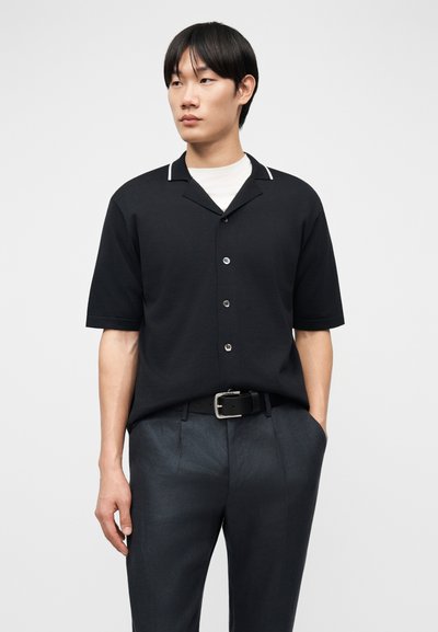 Homme portant une chemise noire à manches courtes boutonnée par-dessus un sous-t-shirt blanc, un pantalon foncé avec une ceinture noire, debout devant un fond uni.