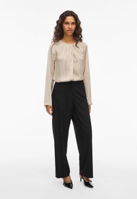 Μπεζ μπλούζα με μακριά μανίκια και πτυχώσεις, συνδυασμένη με μαύρο παντελόνι νάυλον με ψηλή μέση και φαρδιά πόδια, και μαύρες γόβες.