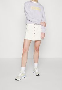 Sweat-shirt violet clair avec le logo "LEVI'S" jaune, associé à une mini-jupe blanche à boutons avant et des baskets de couleur neutre.
