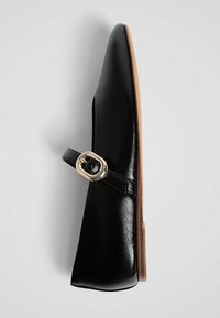 Chaussure plate en cuir verni noir avec une bride unique et une boucle dorée, semelle marron clair, vue de dessus sur fond uni.