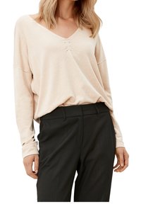 Beige, weicher Strickpullover mit V-Ausschnitt und drei Biesen-Details auf der Brust, kombiniert mit schwarzen, maßgeschneiderten Hosen.