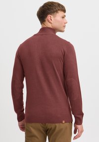 Un pull en maille bordeaux avec un col montant, des poignets et un ourlet côtelés, ainsi que des écussons au niveau des coudes. Il présente une petite étiquette logo en bas.