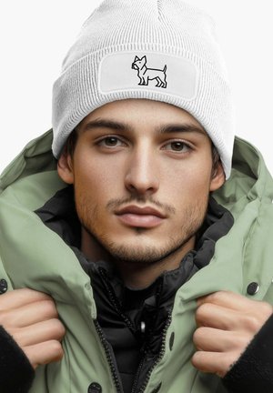 White knitted beanie with a dog graphic on a rectangular patch, worn with a green puffer jacket, featuring black accents and fasteners.