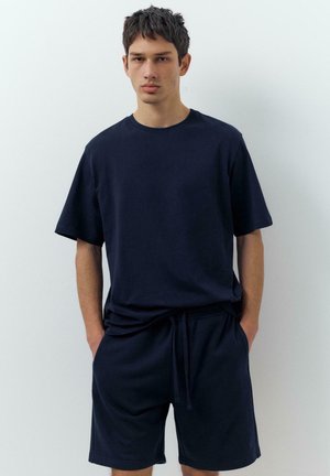 Tânăr stând sprijinit de un perete alb, purtând un tricou larg bleumarin și pantaloni scurți asortați, cu mâinile în buzunare, expresie neutră.