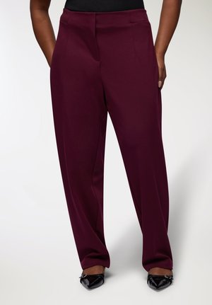 Pantaloni larghi color bordeaux con una texture liscia, dotati di tasche frontali e una vestibilità su misura. Indossati con scarpe nere a punta.