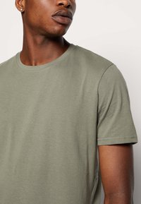 Tricou verde măsliniu cu guler rotund, realizat dintr-un material moale și ușor. Are mâneci scurte, cu o textură netedă și fără modele sau accente vizibile.