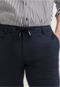 Granatowe dresowe spodnie z elastycznym pasem ze sznurkiem, gładka faktura, kieszenie boczne i detale przeszycia. Noszone z koszulą w paski z guzikami.