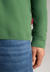 Groen textuurtrui met een ronde halslijn en geribbelde manchetten, gecombineerd met lichtblauwe jeans. Rode merketiket zichtbaar aan de linkerkant.