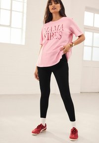 T-shirt in cotone rosa con la scritta "MAMA VIBES", abbinato a leggings neri e scarpe da ginnastica rosse. Calzini bianchi visibili, abbigliamento casual.