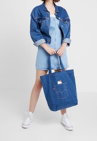 Sac fourre-tout en jean avec une poche avant arborant un motif Hello Kitty, des poign ées robustes et une texture générale en denim bleu.