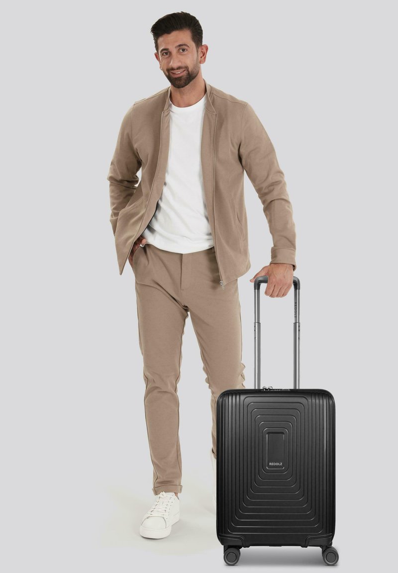 Uomo in abito beige e sneakers bianche è in piedi reggendo il manico di una valigia nera con ruote su uno sfondo grigio.