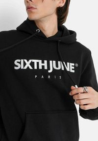 Felpa nera con cappuccio e cordino, caratterizzata da audace scritta bianca "SIXTH JUNE PARIS". Tessuto morbido con tasca frontale e polsini a coste. Anello d'argento alla mano.