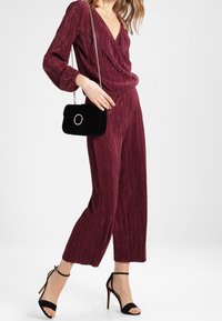 Burgundy, rutigt byxdress med V-ringning och långa ärmar; kombinerat med en liten svart sammetväska och svarta stiletthöga klackar.