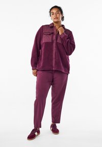 Fleece košile s bundou v tmavě fialové barvě s prošívanou náprsní kapsou, v kombinaci s hladkými fialovými kalhotami a odpovídajícími teniskami.