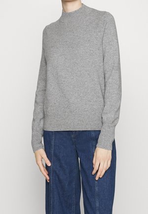 Person iført en ensfarvet lysegrå langærmet sweater og mørkeblå højtaljede jeans, stående med armene afslappet langs siden.
