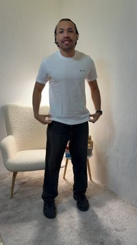 Άνδρας που στέκεται μέσα σε εσωτερικό χώρο φορώντας λευκό μπλουζάκι, σκούρο τζιν και μαύρα παπούτσια, ρυθμίζοντας το πουκάμισό του σε ένα δωμάτιο με μοκέτα, καρέκλα και τραπεζάκι.