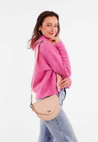 Hnědá kožená crossbody taška s zakřiveným klopou a embosovaným logem, v kombinaci s texturovaným růžovým svetr a modrými džíny. Popruh je nastavitelný.