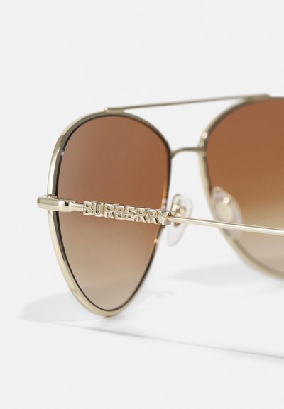 Occhiali da sole in metallo color oro con lenti sfumate marroni. Presentano un prominente marchio "Burberry" sul lato e un design del telaio sottile e elegante.