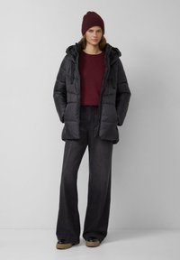 Czarna kurtka puchowa z kapturem, bordowy sweter i czarne spodnie o szerokich nogawkach. Całość dopełniają czarne buty z brązową podeszwą.