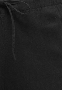 Gros plan sur un pantalon en tissu noir avec un cordon de serrage noué à la taille et des détails de coutures visibles.