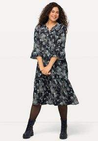 Lang schwarzes Kleid mit Blumenmuster in Hellgrün und Weiß. Besitzt einen Kragen, Dreiviertelärmeln und einen gestuften Rock. Kombiniert mit schwarzen Schnürstiefeln.