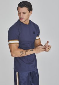 Camiseta de manga corta azul marino con franjas blancas y doradas en las mangas, con un pequeño logo en el pecho. Textura de tela suave.